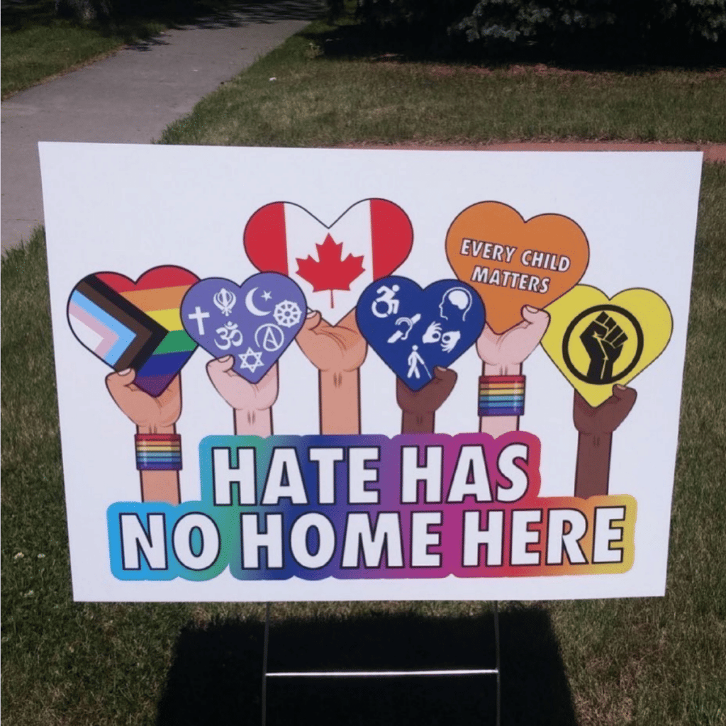 An image of a lawn sign. It’s white, and has in rainbow print Hate has no home here, with various symbols of inclusion and love.