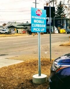 a blue sign is attached to a metal pole that is planted in a cement cylinder, the sign says DQ - reserved for mobile curbside pick-up.